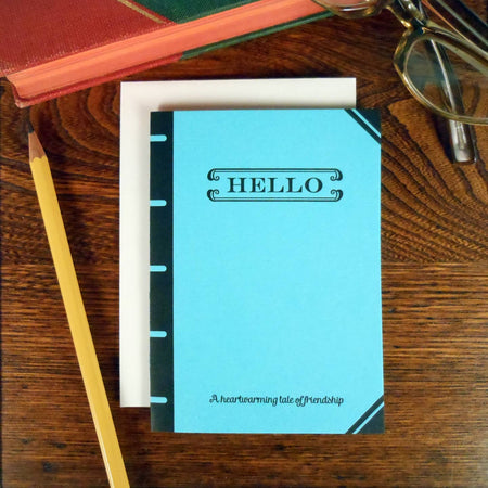 Teal blue card in the image of a vintage book cover with black text saying, “Hello A Heartwarming Tale of Friendship”. A white envelope is included.