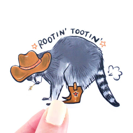 Sticker in the image of a gray raccoon dressed in a cowboy hat and cowboy boots with a cloud of smoke coming out of its tail. Black text saying, “Rootin Tootin”.