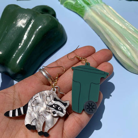 Set of earrings in the image of a gray raccoon and a green trash can.
