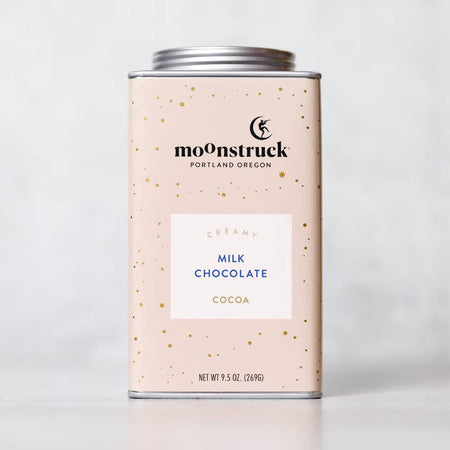 Light pink tin with silver lid. Black text saying, “Moonstruck” with image of a person standing on a half moon. White box in center with blue text saying, “Creamy Milk Chocolate Golden Milk Cocoa”.