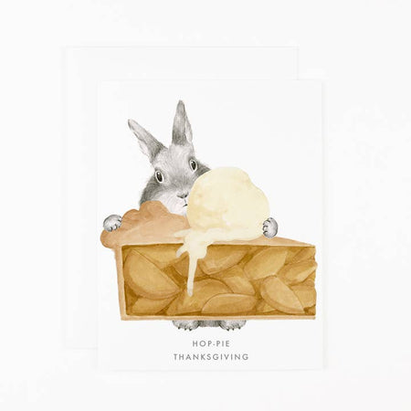 White card with image of a gray bunny holding a piece of apple pie with a scoop of ice cream on top. Gray text saying, “Hop-Pie Thanksgiving”. A white envelope is included.