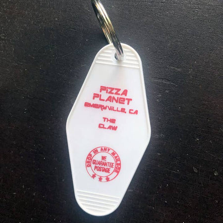 White vintage style motel key fob with red text featuring Pizza Planet and 'the claw' from Toy Story.