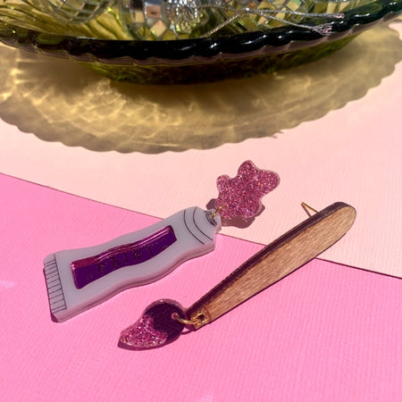 Set of earrings in the image of a brown paintbrush and a white tube of pink paint.