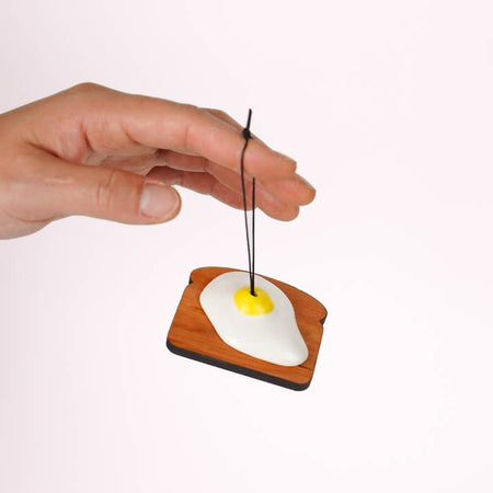 Black string ornament in the image of a wooden piece of toast with a sunny side up egg on it. 