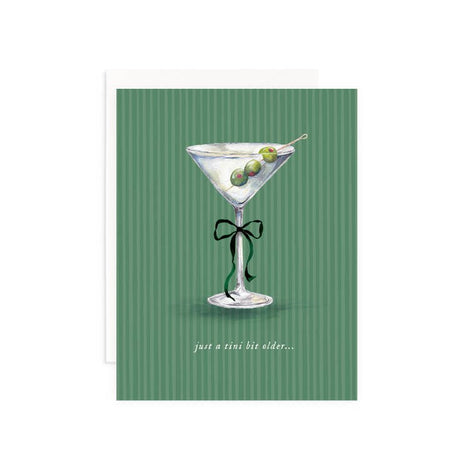 Light green card with dark green vertical stripes and white text saying, “Just A Tini Bit Older…”. Image of a martini drink with green olives on a toothpick and a black bow tied around the stem of the glass. A white envelope is included.