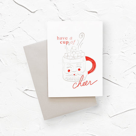 White card with image of a Santa Claus mug with hot chocolate and marshmallows. Red text saying, “Have a Cup of Cheer”. A gray envelope is included.