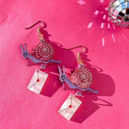 Set of earrings in the image of a pink and purple snail holding a white envelope with a red heart in center.