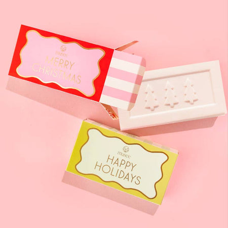 White bar soap with images of embossed Christmas trees in center. Wrapped in a yellow and white box with gold foil text saying, “Happy Holidays”.