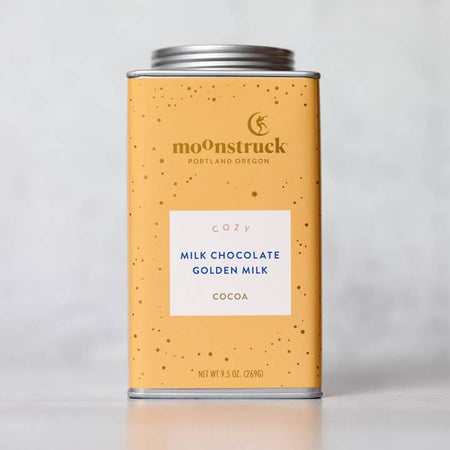 Yellow tin with silver lid. Gold text saying, “Moonstruck” with image of a person standing on a half moon. White box in center with blue text saying, “Cozy Milk Chocolate Golden Milk Cocoa”.