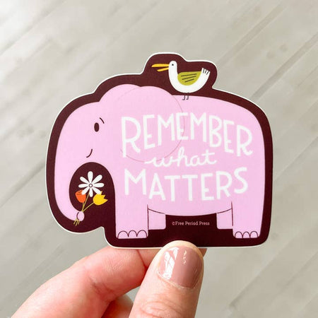 Sticker in the image of a purple elephant holding a bouquet of flowers in her trunk and white bird sitting on her back. White text saying, “Remember What Matters”.