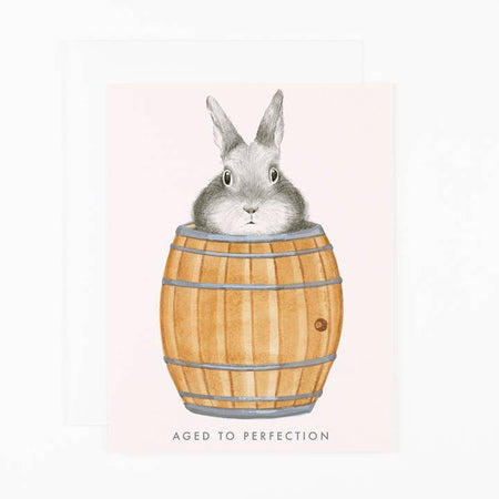 Light pink card with image of a gray bunny inside of a wine barrel. Gray text saying, “Aged to Perfection”. A matching envelope is included.