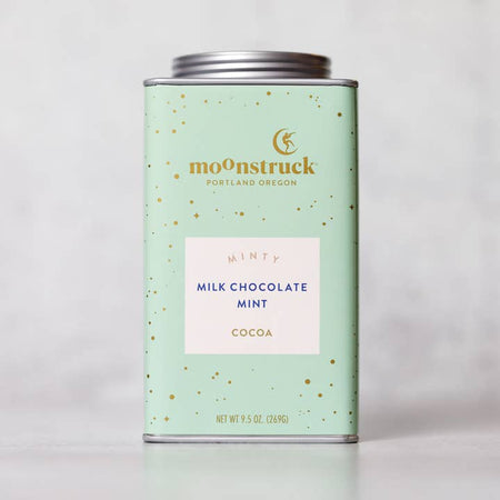 Light green tin with silver lid. Gold text saying, “Moonstruck” with image of a person standing on a half moon. White box in center with blue text saying, “Minty Milk Chocolate Mint Cocoa”.