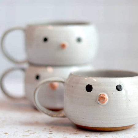 White mug in the image of a snowman. Two black circle eyes and orange triangle nose painted on.