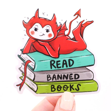 Sticker in the image of a red devil sitting on top of a pile of books with black text saying, “Read Banned Books”.