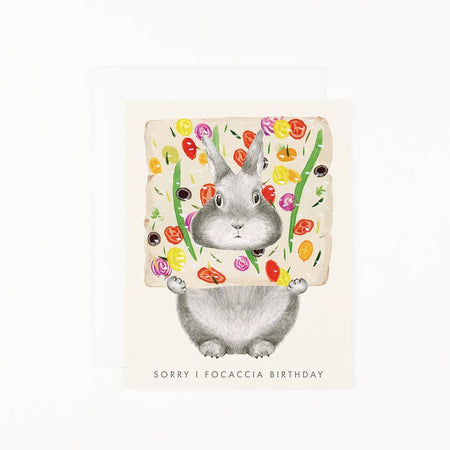 White card with image of a gray bunny holding piece of vegetable focaccia bread. Gray text saying, “Sorry I Focaccia Birthday”. A white envelope is included.