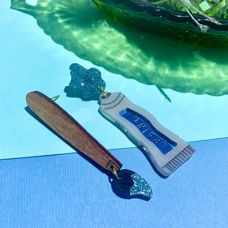 Set of earrings in the image of a brown paintbrush and a white tube of blue paint.