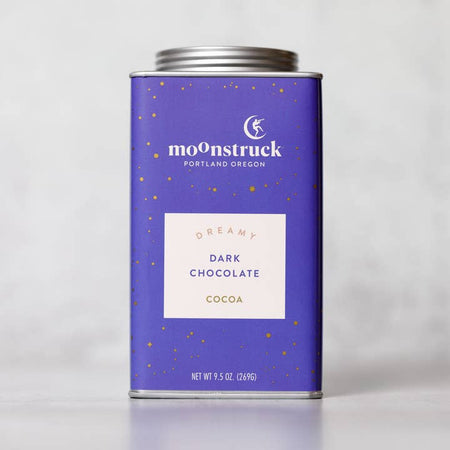 Purple tin with silver lid. White text saying, “Moonstruck” with image of a person standing on a half moon. White box in center with blue text saying, “Dreamy Dark Chocolate Milk Cocoa”.