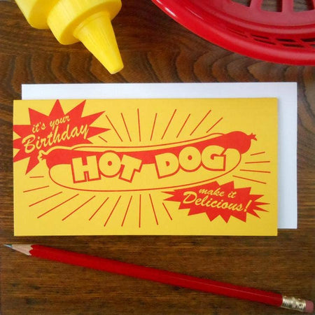 Yellow card with image of a hot dog in a bun. Two red starburst speech bubbles with yellow text saying, “It’s Your Birthday Make It Delicious”.  A white envelope is included.