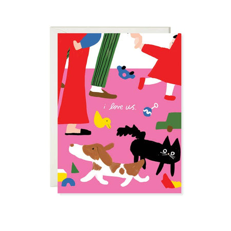 White card with pink carpet with image of a mom pair of legs; a dad pair of legs; a toddler pair of legs; various toys scattered around; and a dog and cat. White text saying, “I Love Us”. A white envelope is included. 