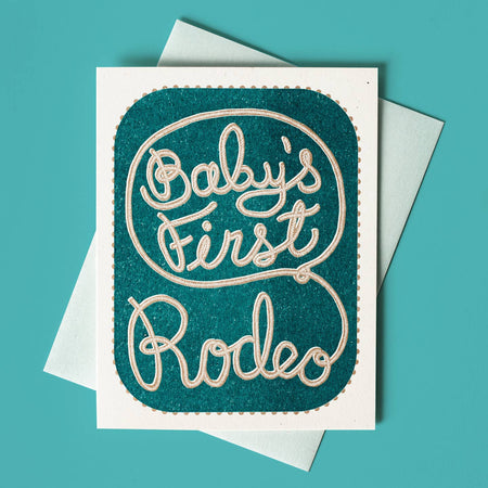 White card with blue insert background and tan text saying, “Baby’s First Rodeo”. Text in the image of rope. A white envelope is included.