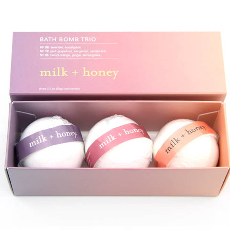 Pink box white text saying, “Bath Bomb Trio” with 3 white bath bombs inside. One with purple ribbon and white text; one with pink ribbon and white text; and one with orange ribbon with white text all saying, “Milk & Honey”.