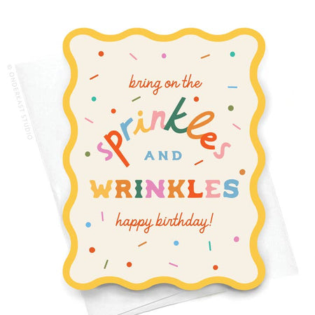 White card with wavy die cut yellow border. Orange and rainbow text saying, “Bring On the Sprinkles and Wrinkles Happy Birthday!” Colored sprinkled scattered across the card. A white envelope is included.