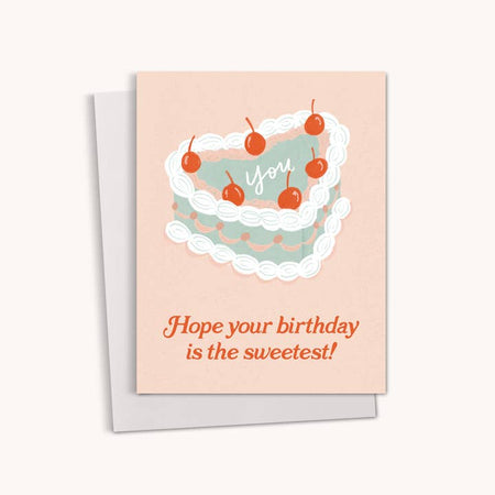 Tan card with red text saying, “Hope Your Birthday is the Sweetest!” Image of a heart shaped cake with green frosting and white border and red cherries on top. White text saying, “YOU” in the middle of the cake. A gray envelope is included.