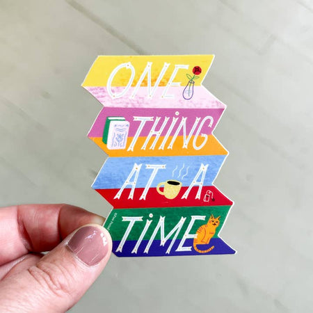 Sticker in the image of a set of stairs that are each a different color with an orange cat, cut of tea, and books. White text saying, “One Thing At A Time”.