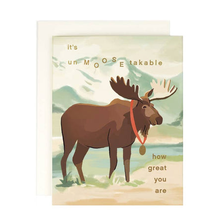 White card with image of a mountain and stream with a brown moose wearing a gold medal in center. Gold foil text saying, 