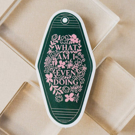 Green sticker in the image of a vintage motel keychain with pink flowers and pink text saying, “What Am I Even Doing”.